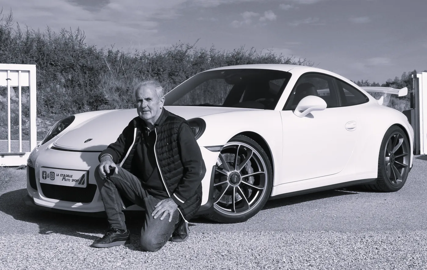 Robert Buisson, fondateur de La Citadelle, un genou à terre devant une Porsche blanche à l’entrée du garage